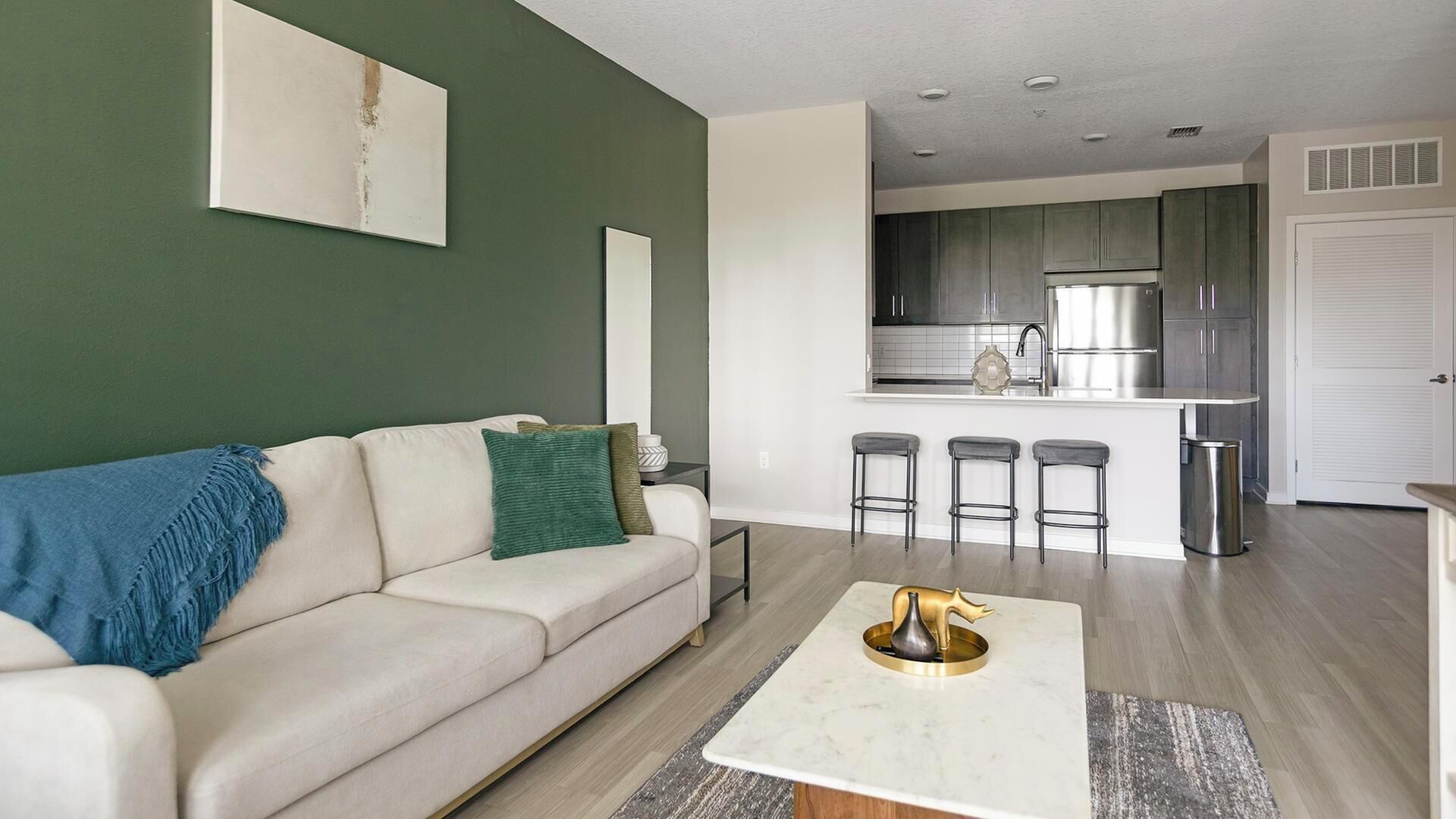 A modern living room with a sofa, a coffee table, and a kitchen area with a refrigerator and bar stools.