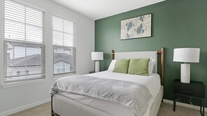 Une chambre à coucher avec un lit, deux tables de chevet, une fenêtre avec des stores et un mur vert orné d’un tableau.
