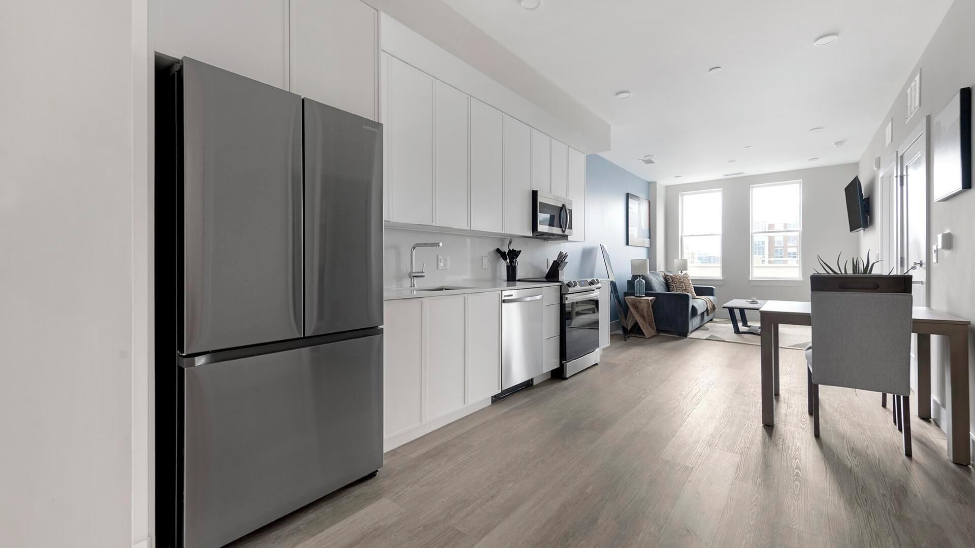 A modern kitchen with white cabinetry, a stainless steel refrigerator, and a dining area with a table and chairs.