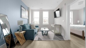 Une chambre moderne avec un canapé bleu, une petite table, un téléviseur fixé au mur et un lit dans la pièce adjacente.