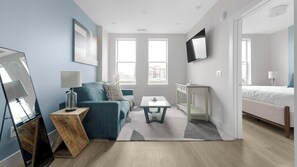Ein modernes Zimmer mit einem blauen Sofa, einem kleinen Tisch, einem an der Wand montierten Fernseher und einem Bett im angrenzenden Raum.