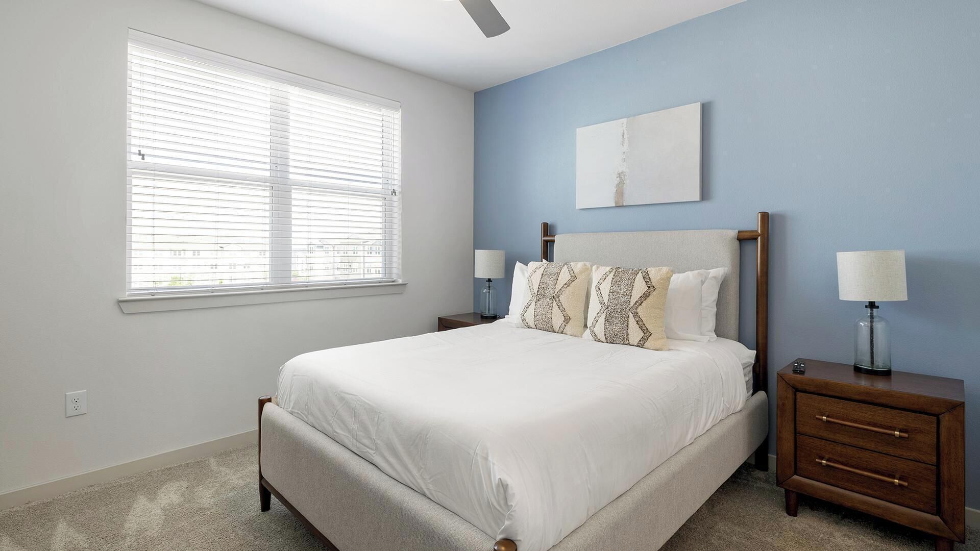 A bedroom with a bed, two nightstands, a ceiling fan, a window with blinds, and a wall-mounted artwork.