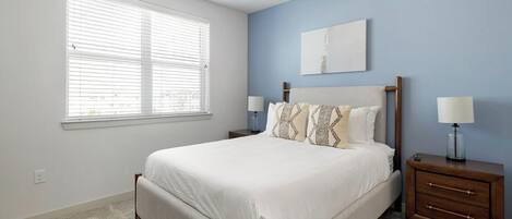 A bedroom with a bed, two nightstands, a ceiling fan, a window with blinds, and a wall-mounted artwork.