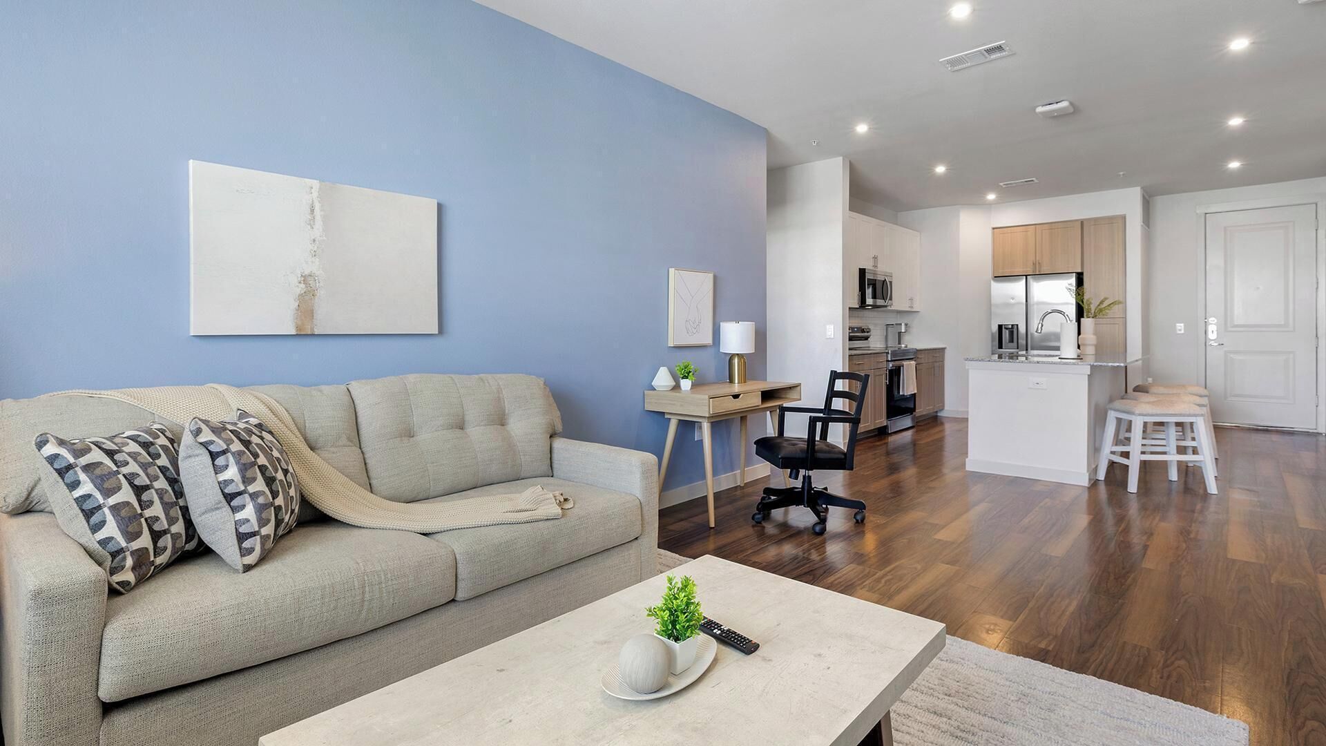 A living room with a sofa, coffee table, and a desk with a lamp. There is a kitchen area with cabinets and a refrigerator.
