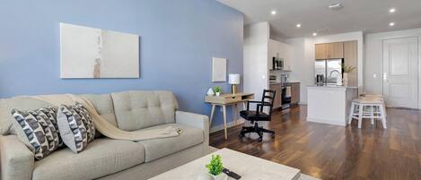 A living room with a sofa, coffee table, and a desk with a lamp. There is a kitchen area with cabinets and a refrigerator.
