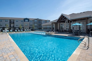 A rectangular swimming pool with a tiled deck, surrounded by lounge chairs and umbrellas, with multi-story buildings in the background. - Landing | Incredible 2BD, Clubhouse, Pool (Rockwall)