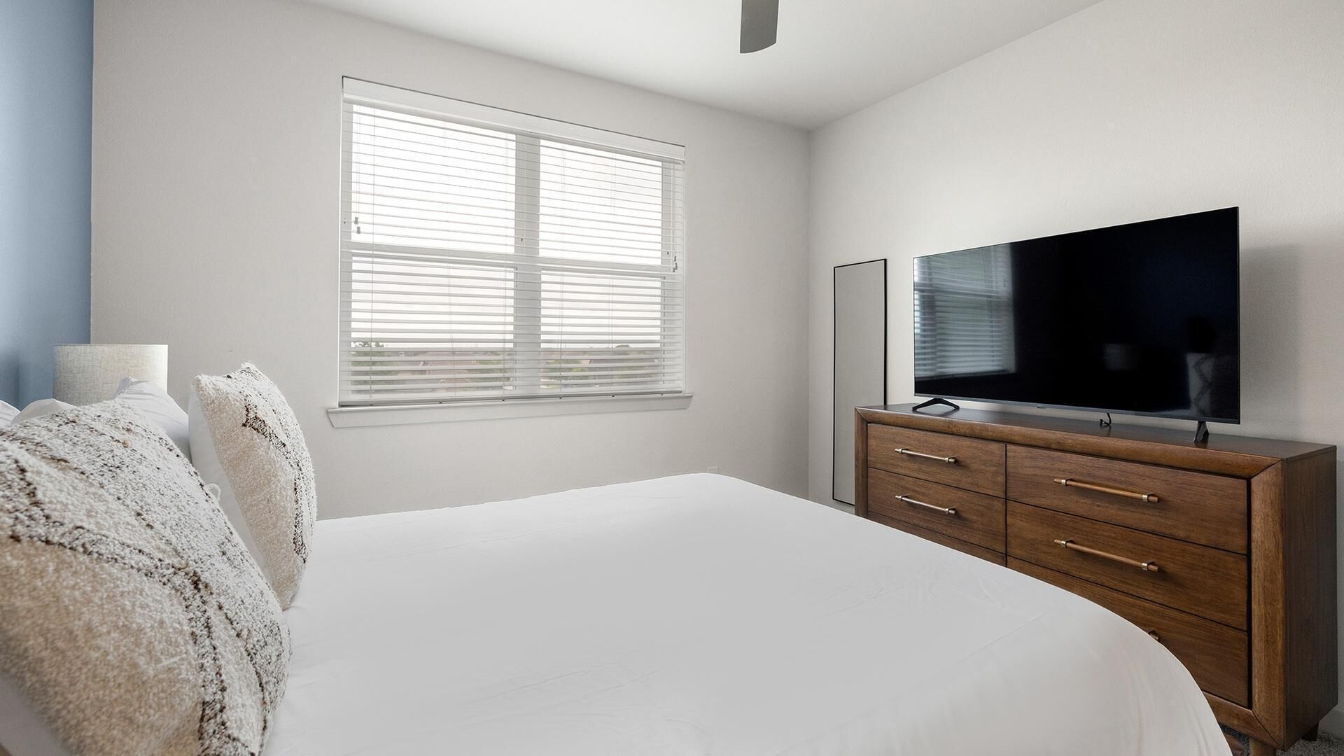 Un dormitorio con cama, cómoda, televisor y ventana con persianas.