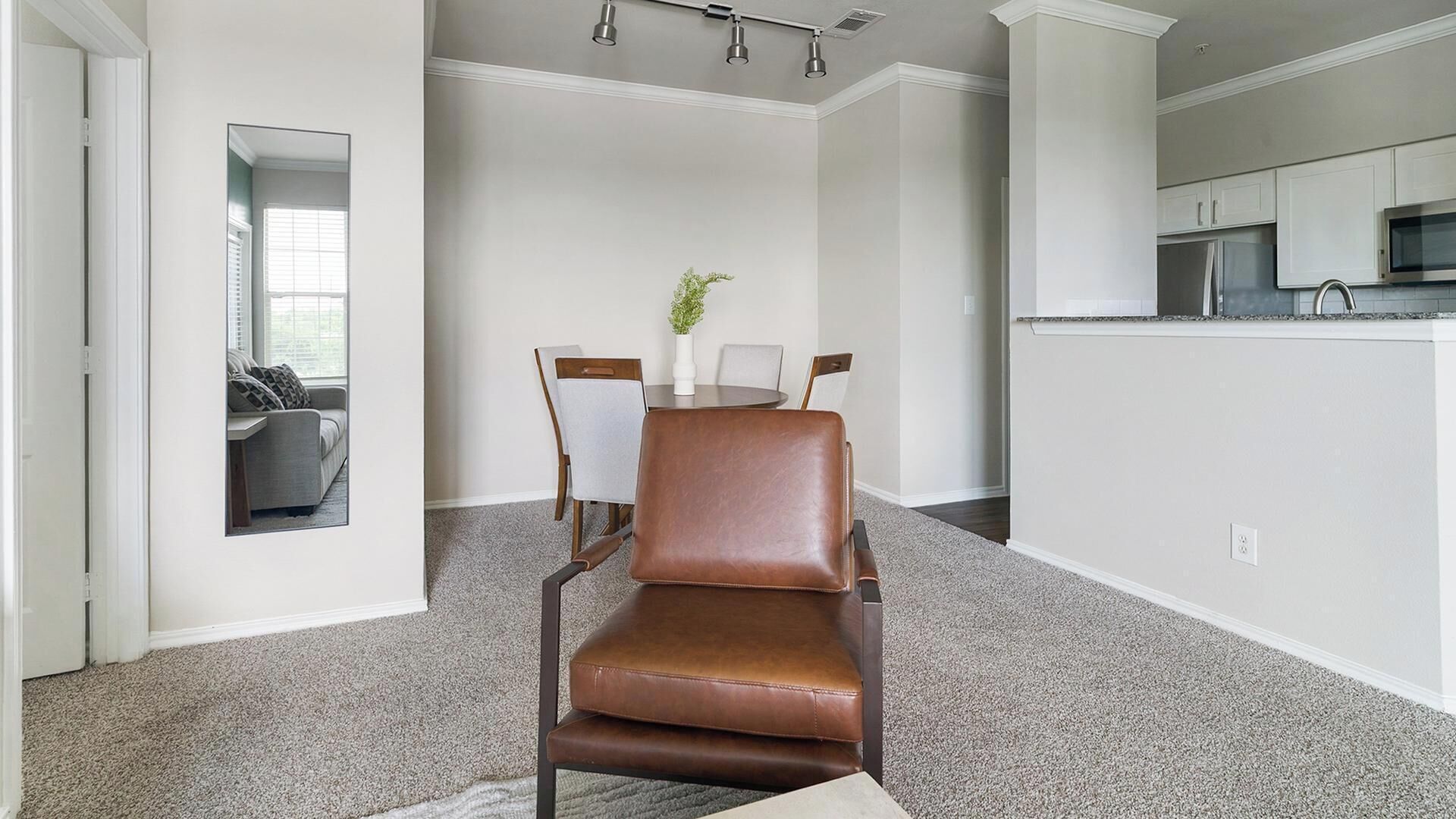 Una sala de estar moderna con un sillón de cuero marrón, una zona de comedor con una mesa y sillas blancas, y una cocina con armarios blancos y un microondas.