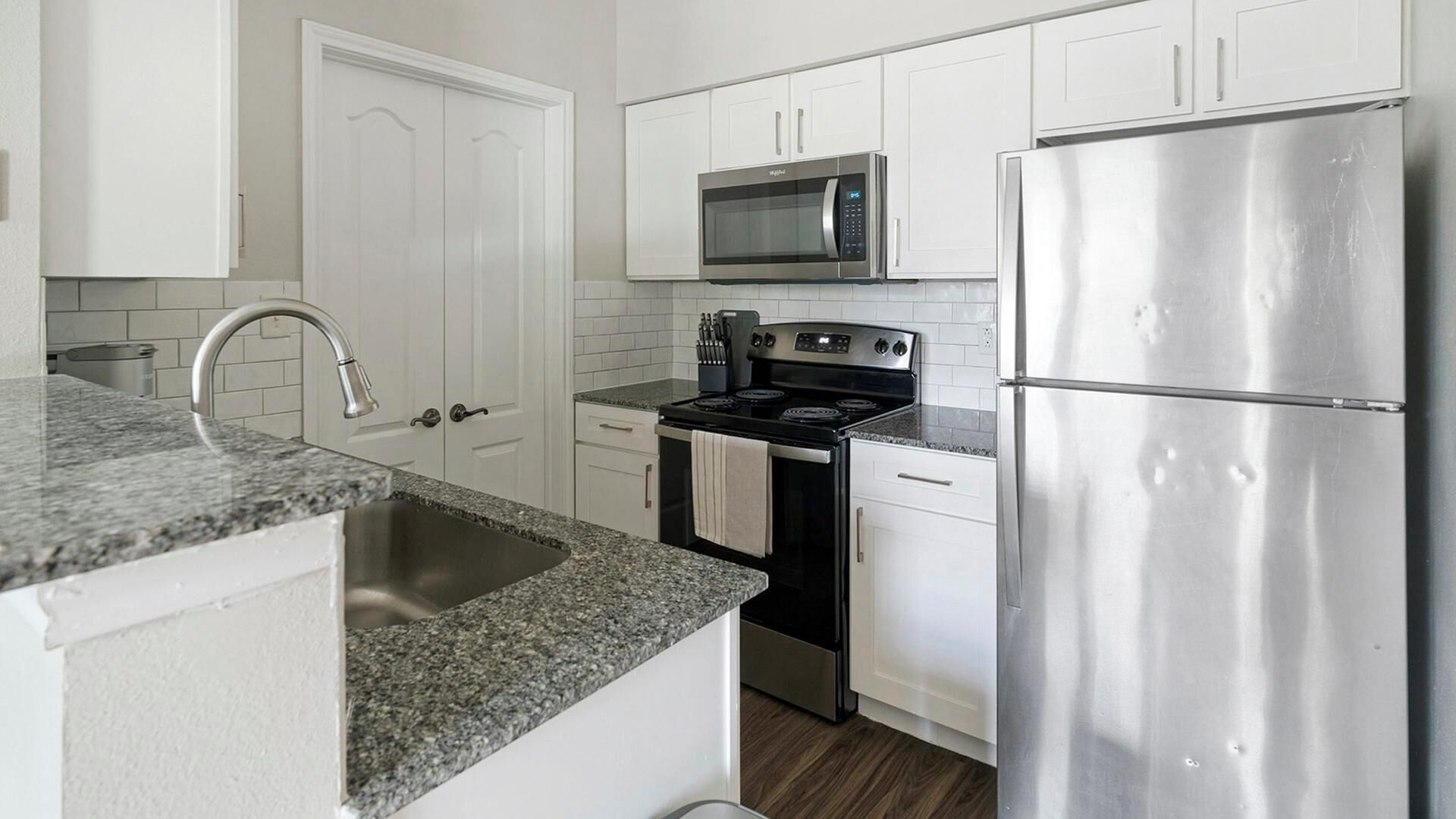 A modern kitchen with white cabinetry, a stainless steel refrigerator, a microwave, an oven, and a granite countertop.