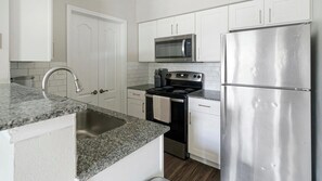 A modern kitchen with white cabinetry, a stainless steel refrigerator, a microwave, an oven, and a granite countertop. - Landing | Stylish 1BD in Copperfield (Cypress)