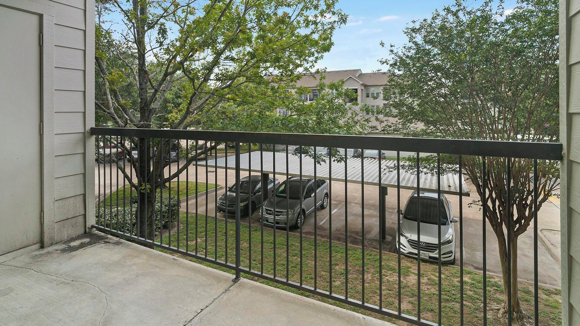 A balcony with a metal railing overlooking a parking area with several cars and trees.