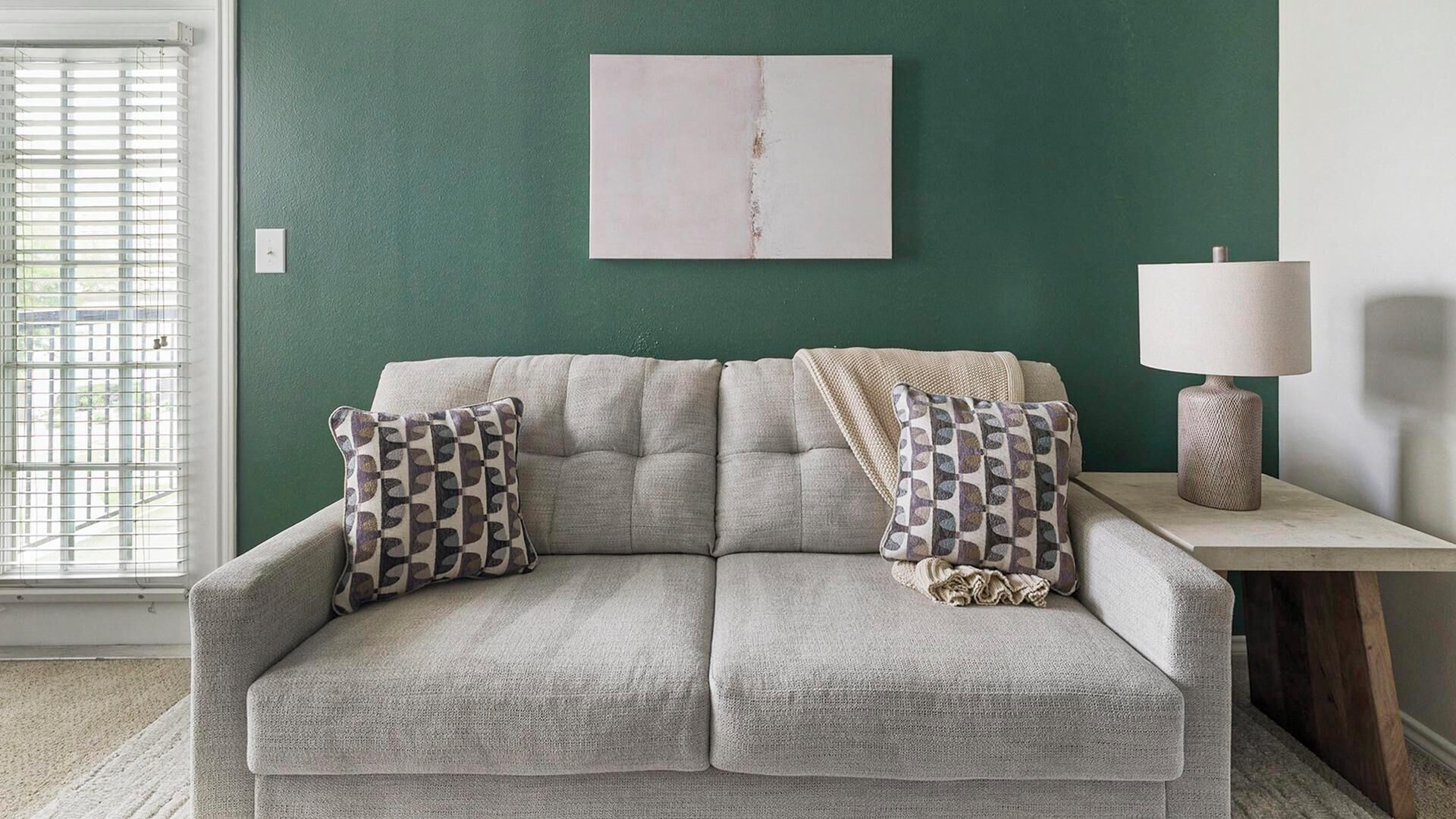 Un canapé agrémenté de coussins à motifs, une lampe posée sur une table d’appoint et un mur orné d’une seule œuvre d’art.