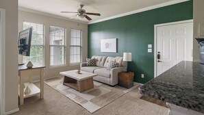Un salon avec un canapé, une table basse et un téléviseur. Il y a des fenêtres avec des stores, un ventilateur de plafond et une porte.