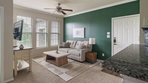 A living room with a sofa, coffee table, and a TV. There are windows with blinds, a ceiling fan, and a door. - Landing | Stylish 1BD in Copperfield (Cypress)