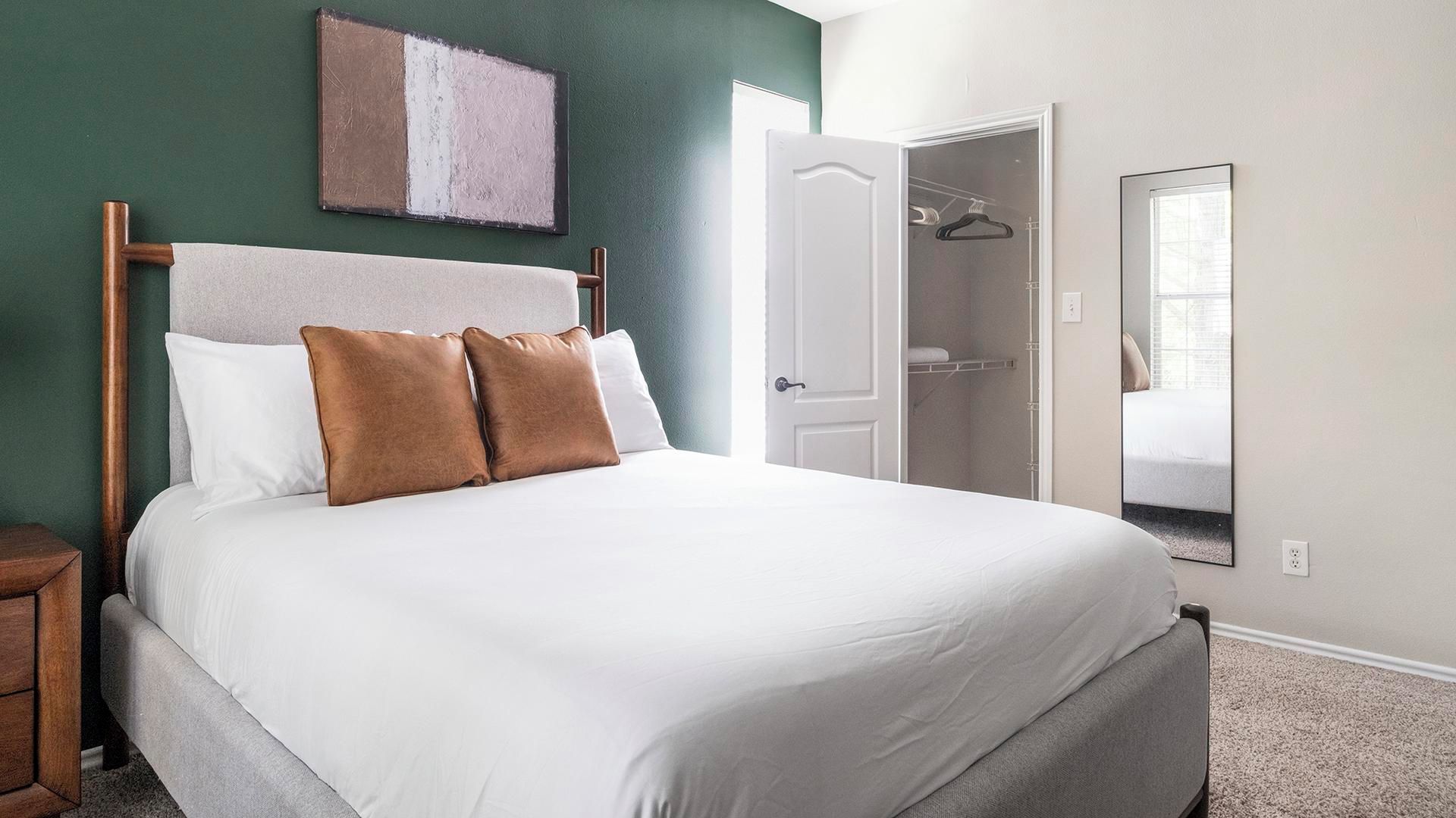 Una cama bien hecha con cabecera, una mesita de noche de madera, un espejo y una puerta que da al baño.