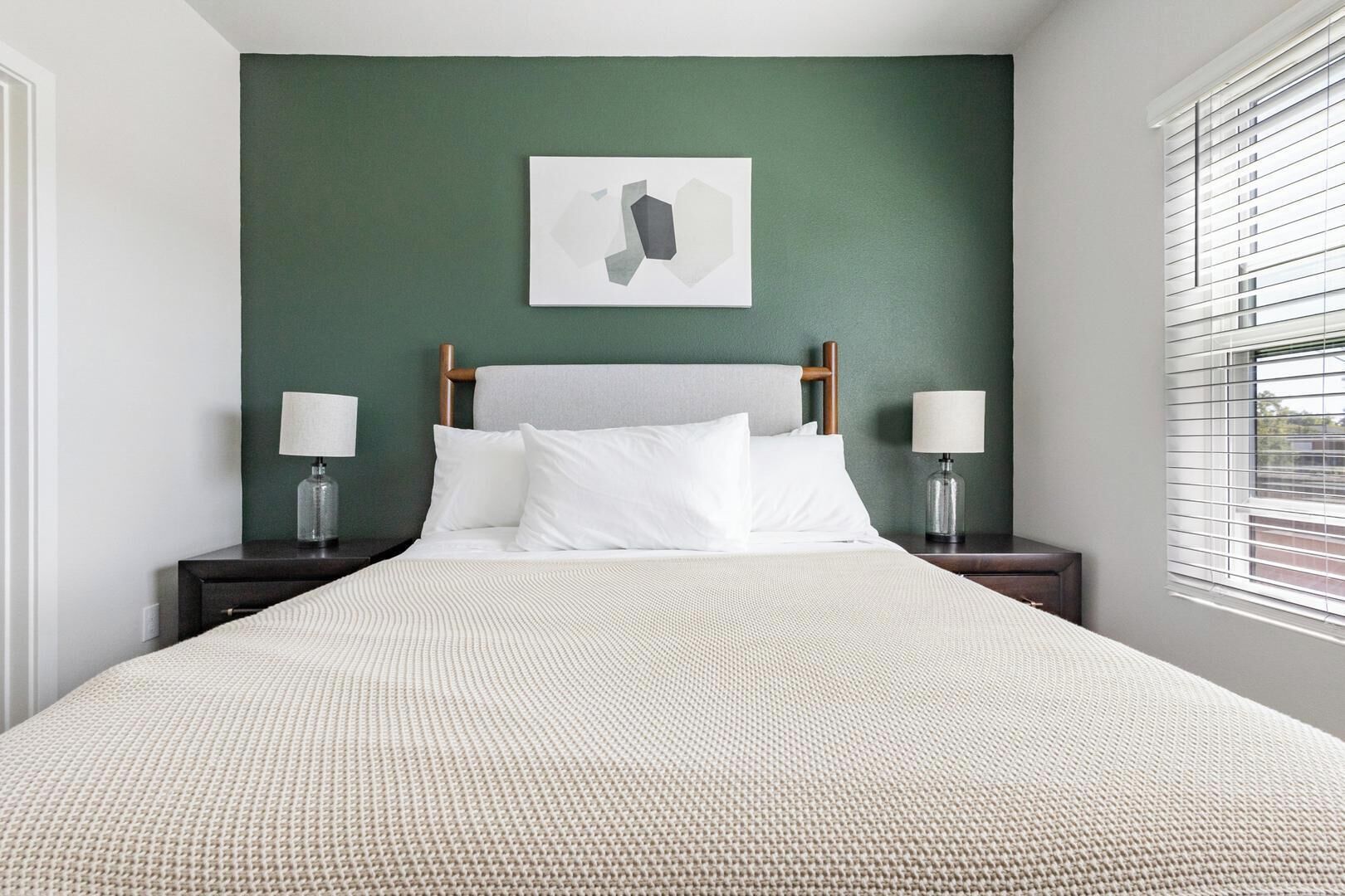 A neatly made bed with a textured white cover, two bedside tables with lamps, a green wall, and a framed picture above the bed.