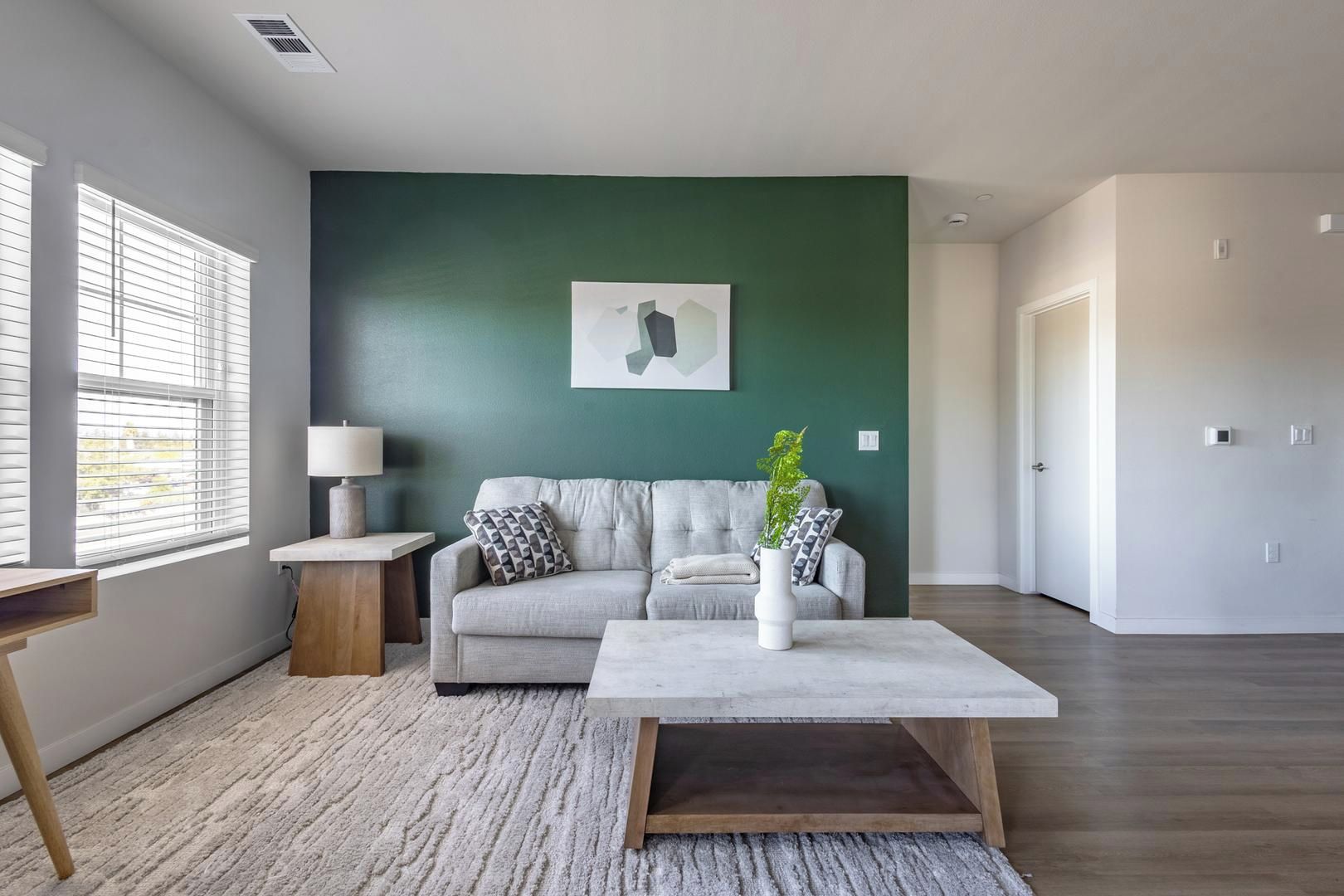Een woonkamer met een grijze bank, een witte salontafel, een lamp en een groene wand.