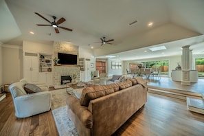 A spacious living room with a stone fireplace, ceiling fans, and a large TV. - Pool, Theater, Game Room + Massive Private Lawn (Frisco)