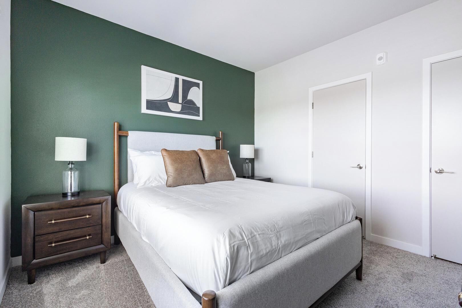 A neatly arranged bedroom with a bed, two bedside tables, a nightstand, a door, and a wall-mounted artwork.