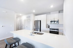 A modern kitchen with white cabinetry, stainless steel appliances, and a central island with bar stools. - Landing | Beautiful 2BD, Courtyard, Gym (Santa Rosa)