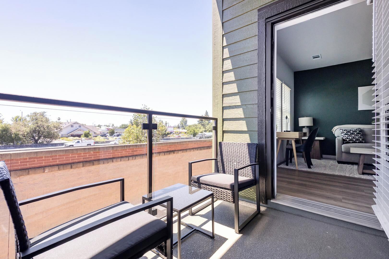 Varanda com vista para um parque de estacionamento e edifícios, uma pequena mesa com uma cadeira, e uma porta que dá para um quarto com um sofá e um candeeiro.