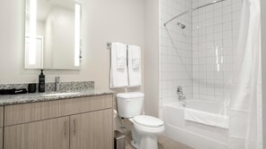A modern bathroom with a granite countertop, a toilet, a sink, a mirror, and a shower with a glass door. - Landing | Stunning 2BD in Downtown (Savannah)