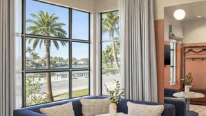 Une chambre d’hôtel avec de grandes fenêtres, un canapé bleu, une petite table ronde et une vue sur des palmiers et une plage.
