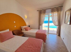 Une chambre d’hôtel avec deux lits, un mur orange incurvé, une vue sur la mer et une grande fenêtre avec des rideaux.