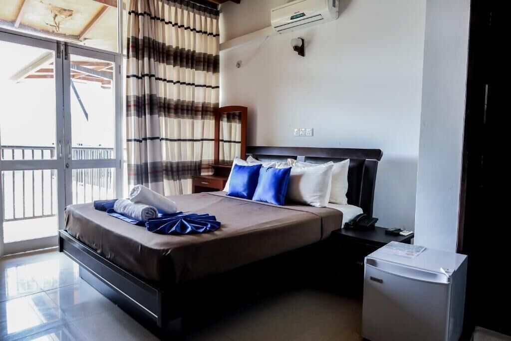 A hotel room with a bed, a small desk, a refrigerator, a window with curtains, and a balcony.