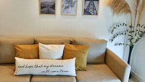 Un sofá color beige con tres cojines, una pared adornada con tres cuadros enmarcados y una planta alta con plumas de pampa.