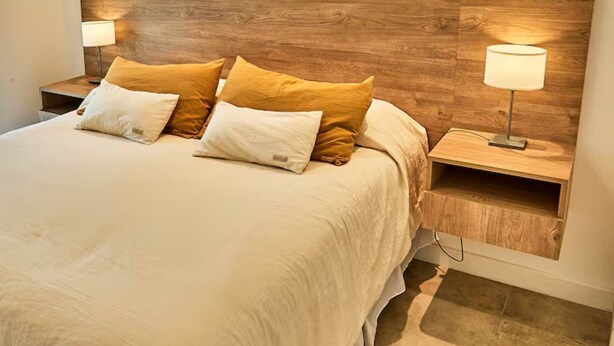 Una cama con cabecera de madera, dos mesitas de noche con lámparas y una colcha en tonos neutros.