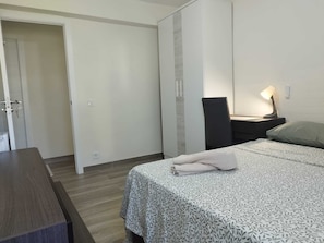 A bedroom with a bed, a desk, a chair, a nightstand, and a door.