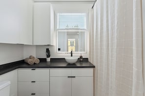 Un baño moderno con un lavamanos blanco, encimera negra y una ventana con persianas.