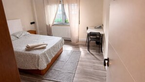 Une chambre à coucher comprenant un lit, un bureau, une chaise, une fenêtre avec des rideaux, un luminaire suspendu et un panneau décoratif mural.