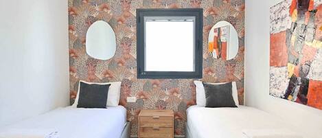 A room with two beds, a small wooden nightstand, a window, and a wall with a floral pattern.