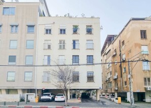 A multi-story building with a parking area in front, featuring a small tree and two parked cars. - Hayarkon Joie - Two Bedroom Apartment, Sleeps 4 (Tel Aviv-Yafo)
