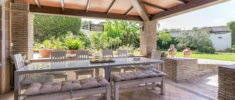 Un patio al aire libre con una mesa de vidrio, bancos acolchados y un pilar de ladrillo.