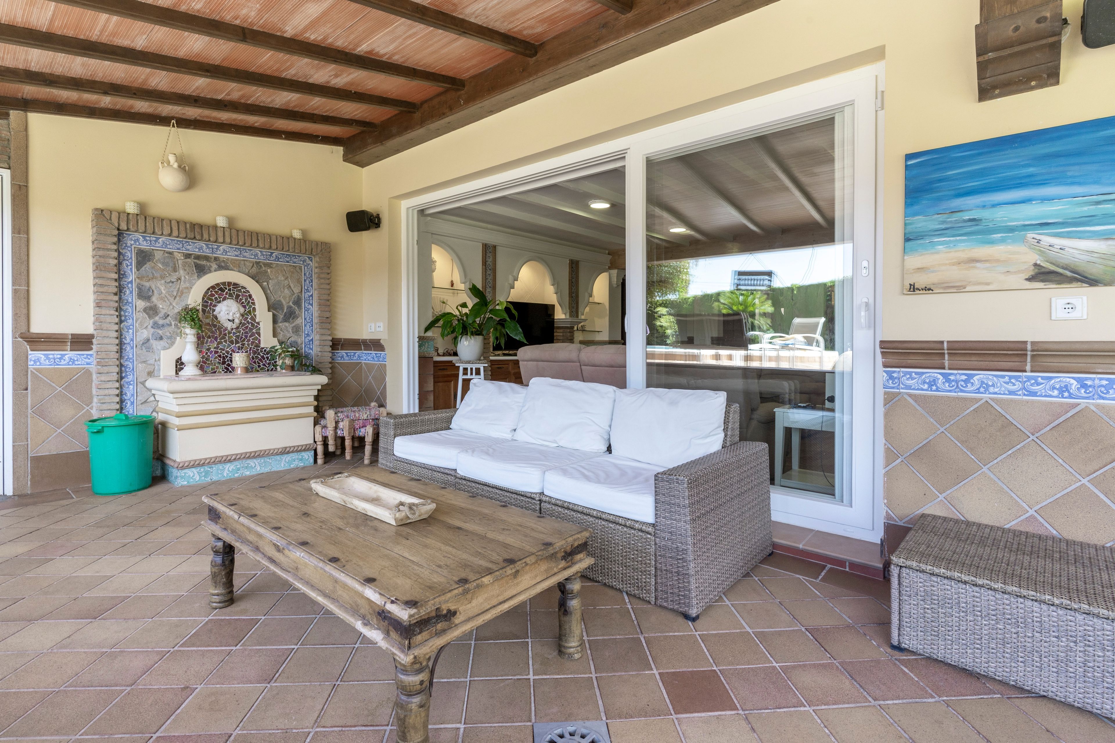 A covered patio area with a wicker sofa, a wooden table, and a tiled floor.