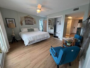 Une chambre d’hôtel équipée d’un lit, d’un bureau, d’une chaise, d’un ventilateur de plafond et donnant sur le couloir.