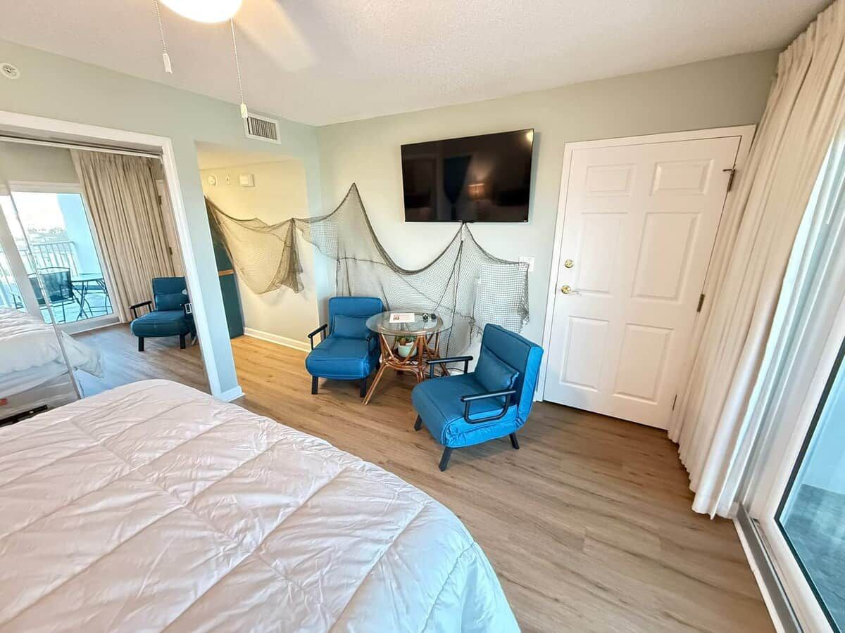 Una habitación de hotel con una cama, dos sillas azules, una mesita, un televisor y una puerta.