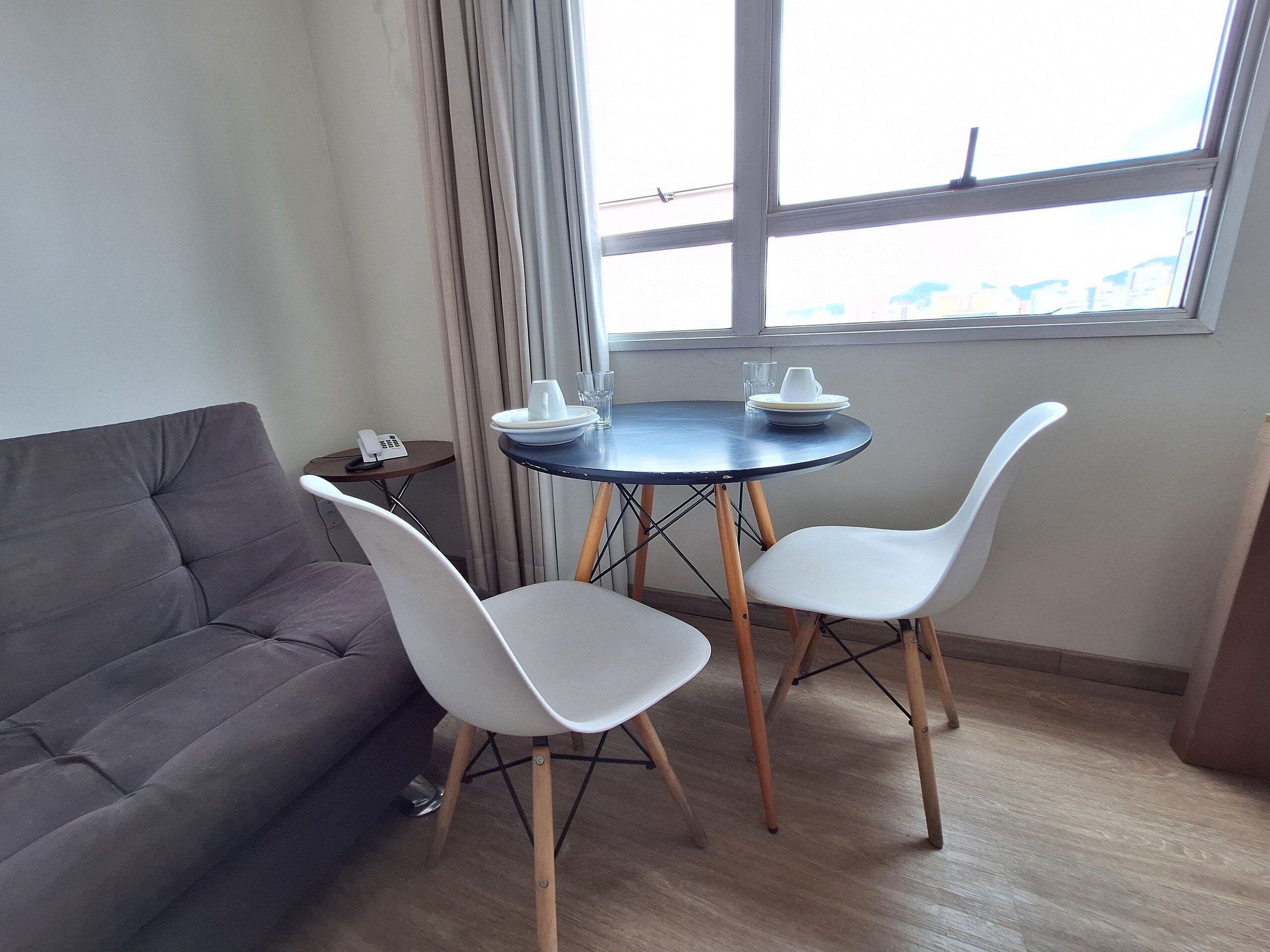 Una habitación pequeña con un sofá gris, una mesa redonda con dos sillas blancas, una mesita auxiliar con un teléfono, y una ventana con vistas a edificios.