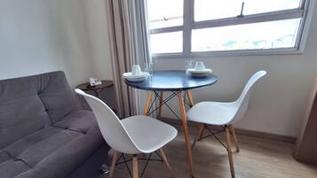 Una habitación pequeña con un sofá gris, una mesa redonda con dos sillas blancas, una mesita auxiliar con un teléfono, y una ventana con vistas a edificios.
