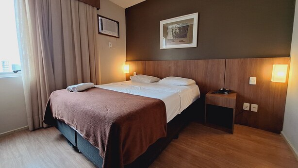 Una habitación de hotel con cama, mesita de noche, lámpara de pared y ventana con cortinas.