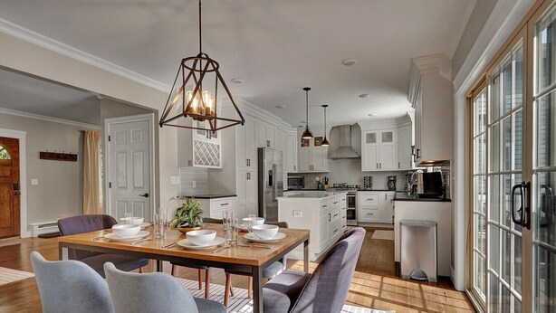 A modern kitchen with white cabinetry, a dining area with a wooden table and grey chairs, and a large window with a view. - Central Royal Oak 3BR Home Near Woodward, Birmingham & Detroit Zoo (Royal Oak)