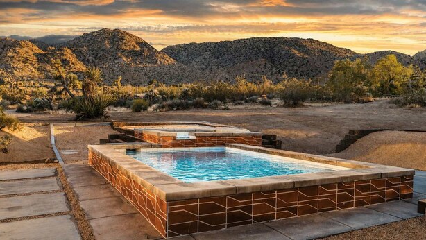 Una piscina con borde de azulejos, rodeada por una terraza de piedra y con montañas al fondo.