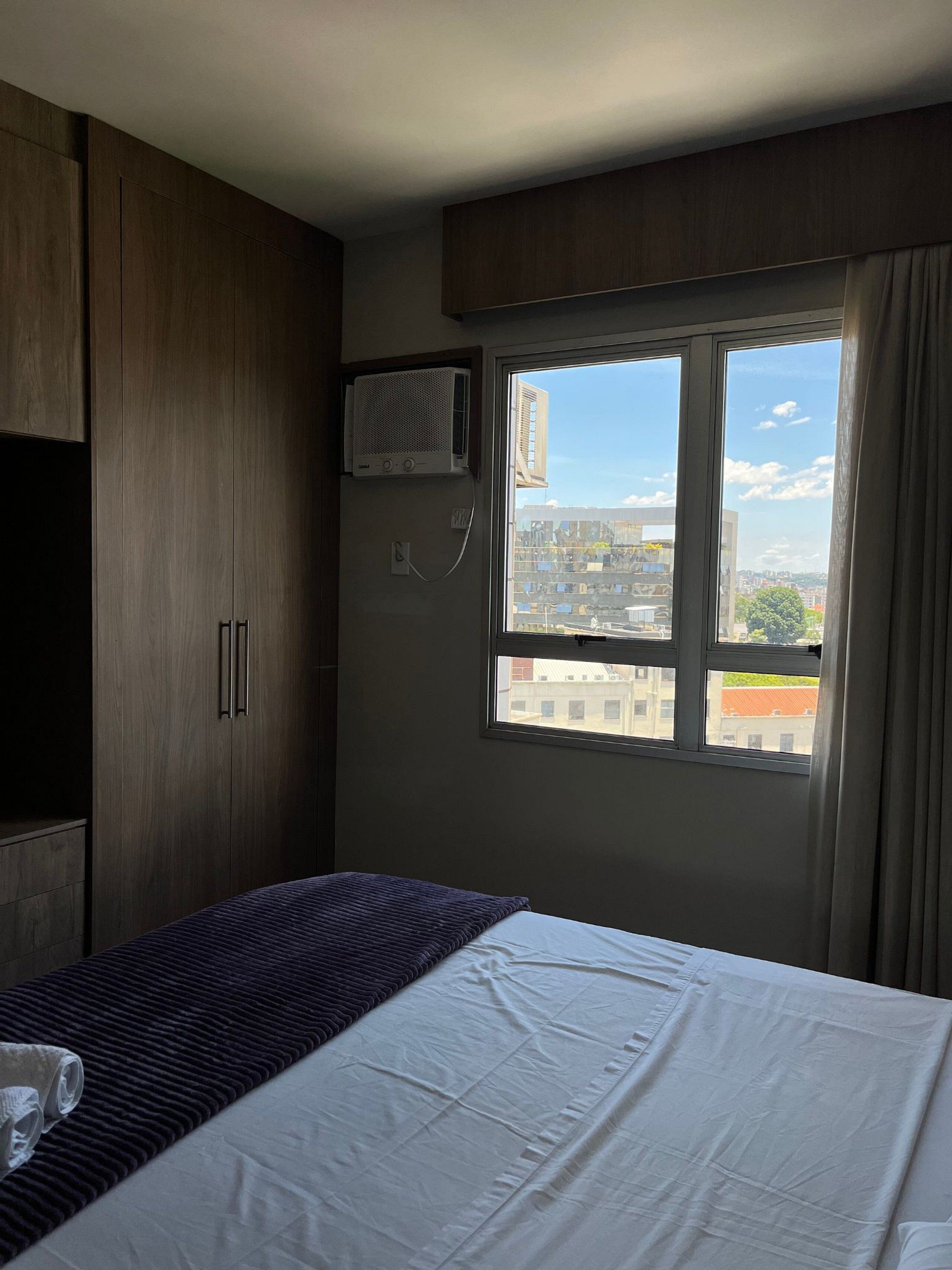 Habitación de hotel con cama, ventana con vista a edificios, armario y unidad de aire acondicionado.