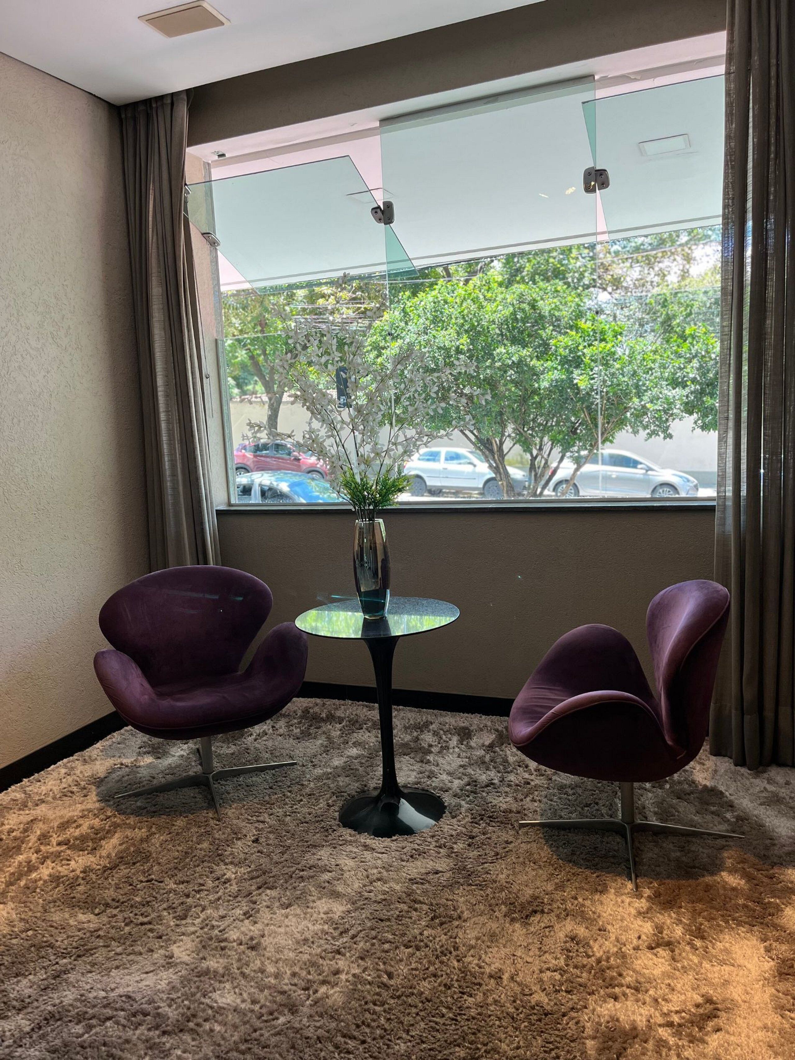 A room with two purple chairs, a small table with a plant, and a view of parked cars and trees through a window.