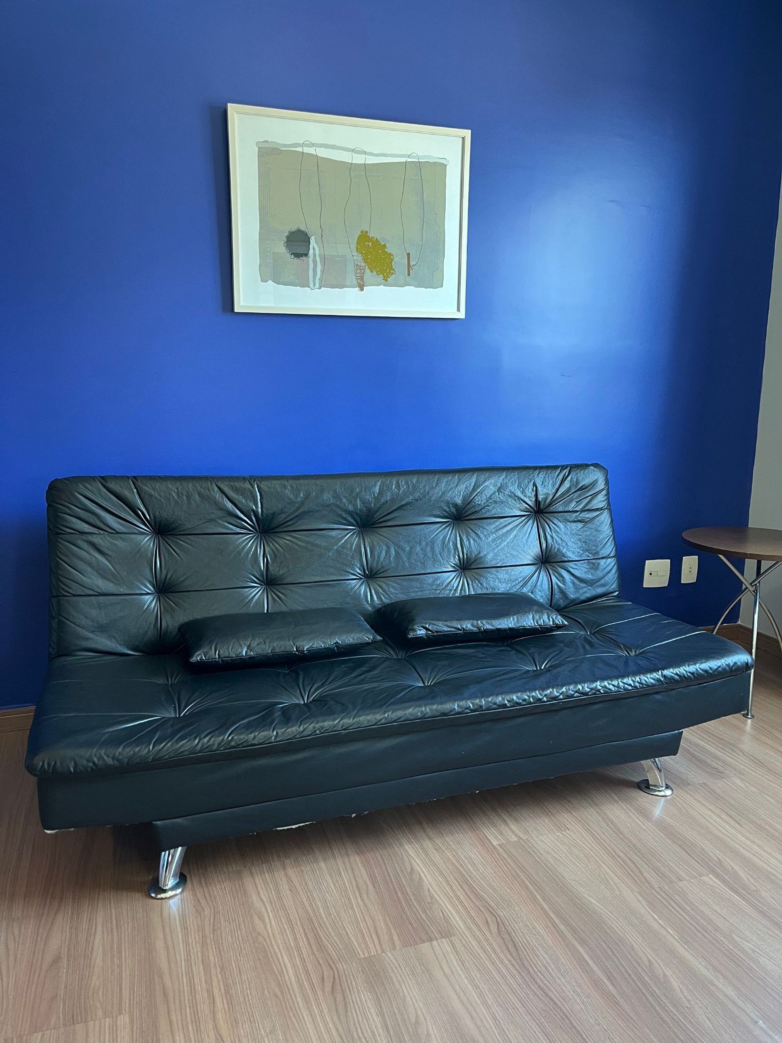 A black leather sofa with a tufted backrest and metal legs, positioned against a blue wall with a framed abstract painting above it.