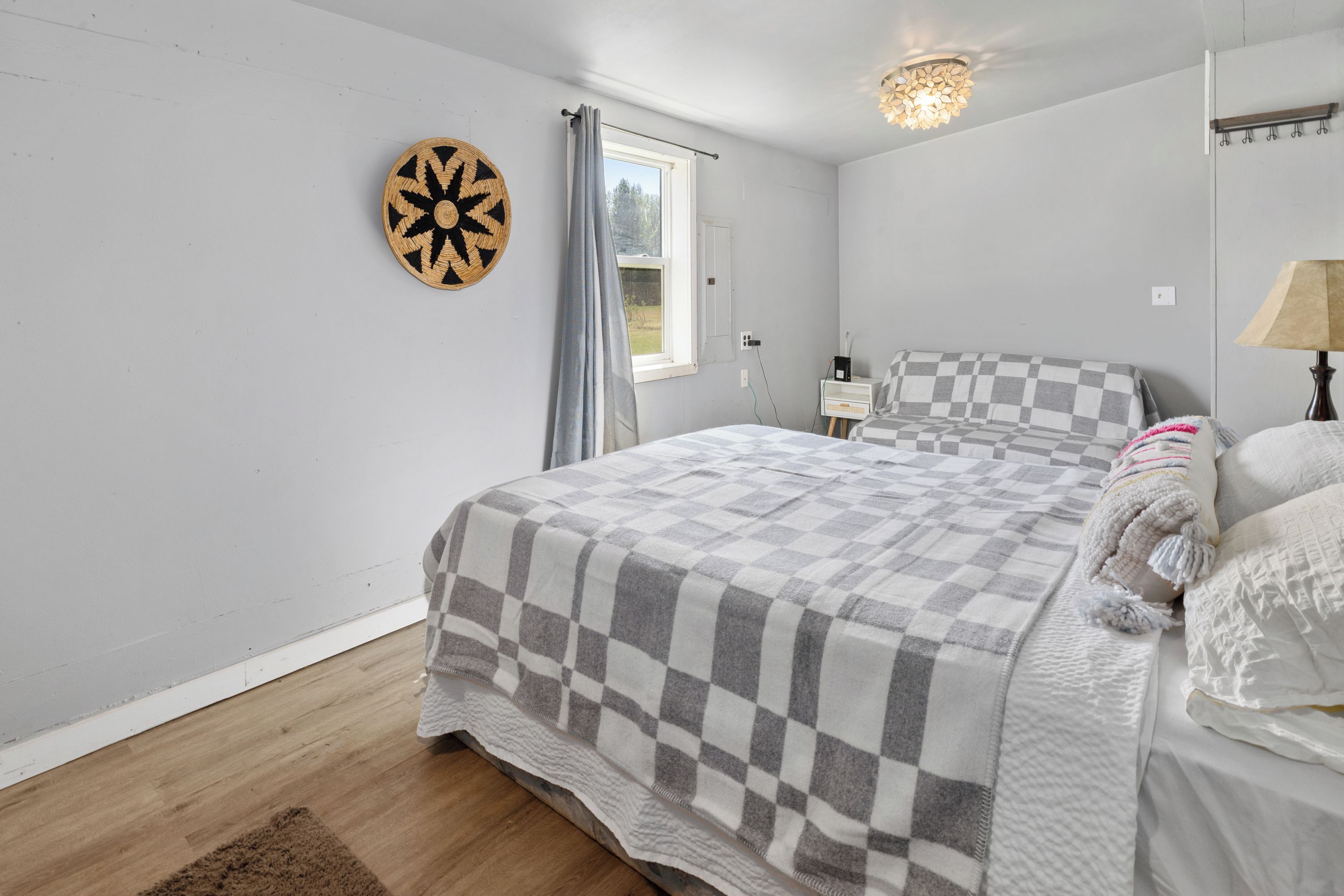 A bedroom with a checkered bedspread, a dartboard, a window with curtains, a bedside table, and a lamp.