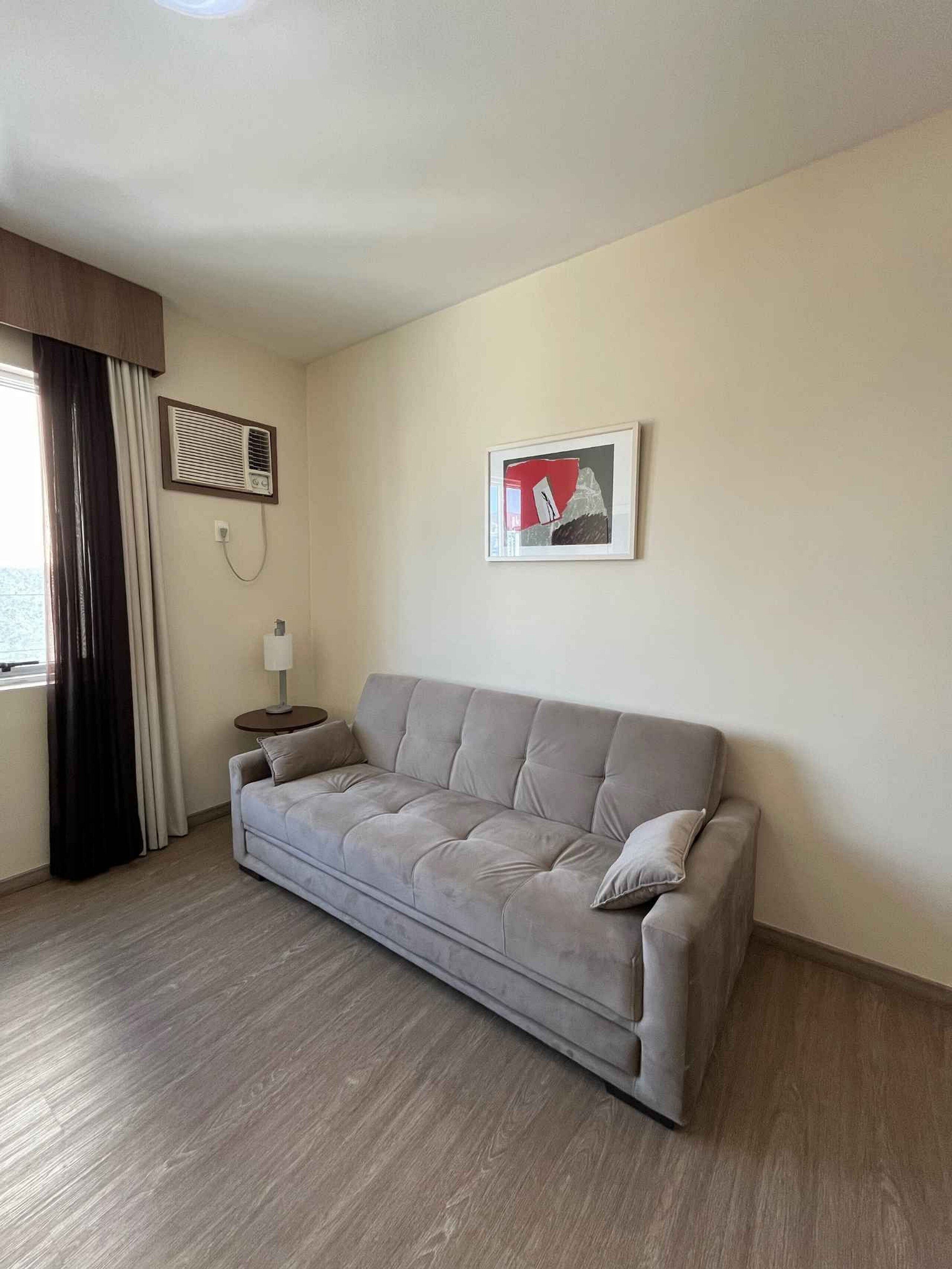 A neutral-toned room with a grey sofa, a small side table, a framed picture on the wall, and a window with curtains.